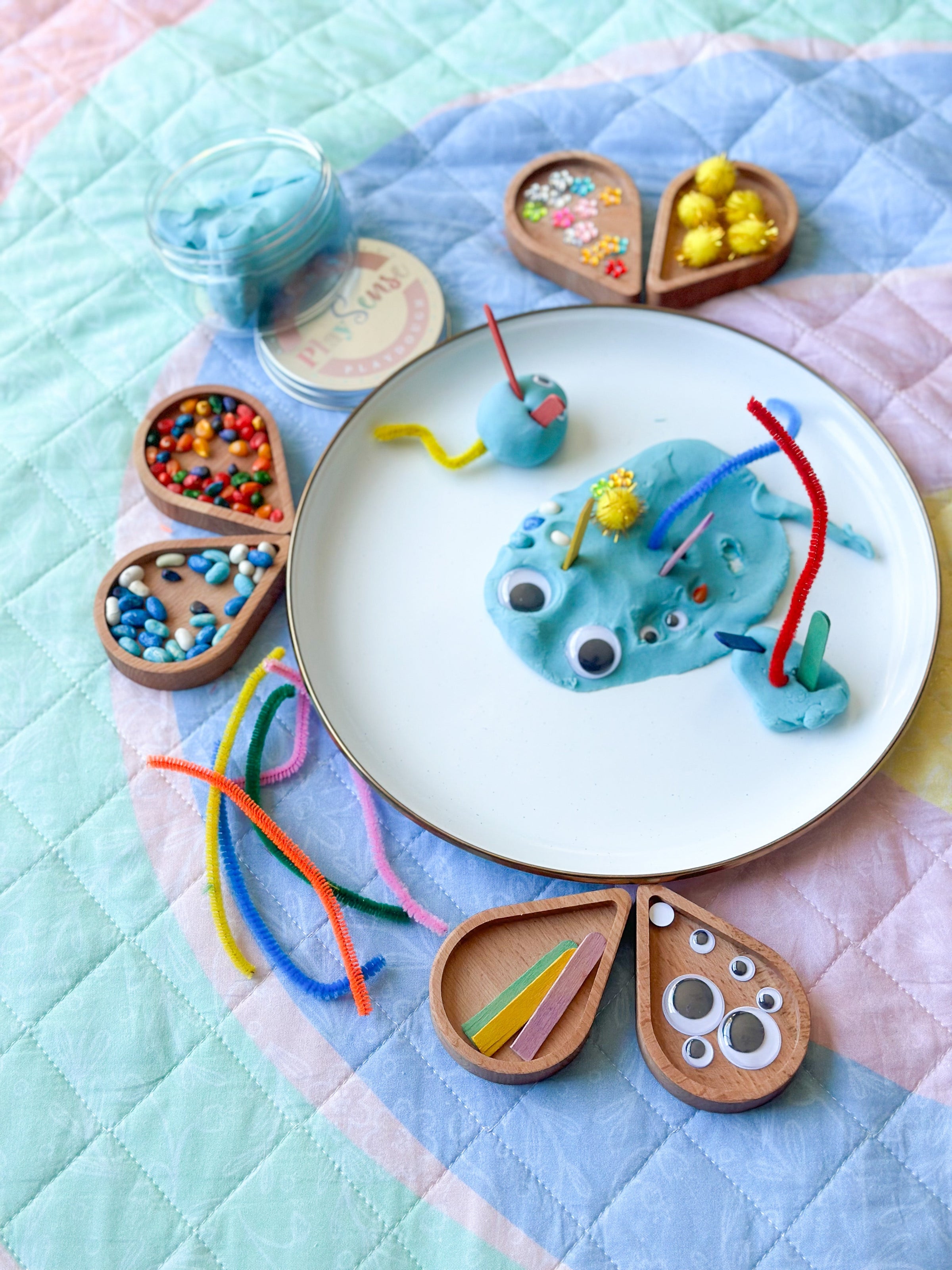 Frogs made from play dough on a plate with colorful sticks and small wooden containers on a quilted fabric background.
